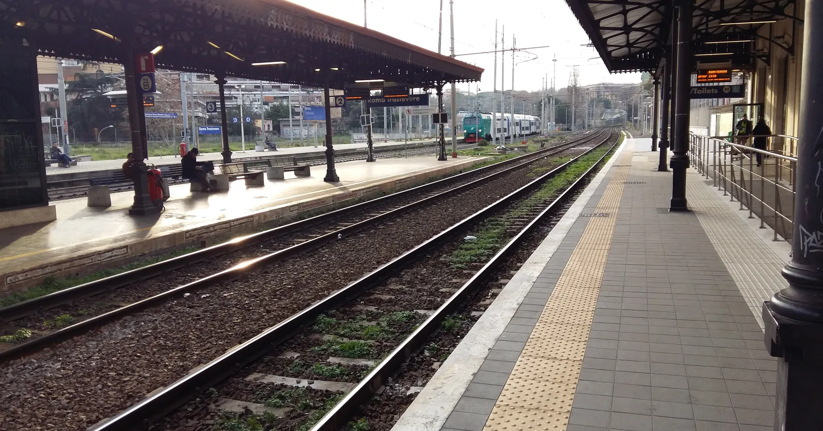 The platforms at Roma Trastevere station under an old iron and glass canopy on cast-iron columns, tracks running away between them with a regional train standing at the far end