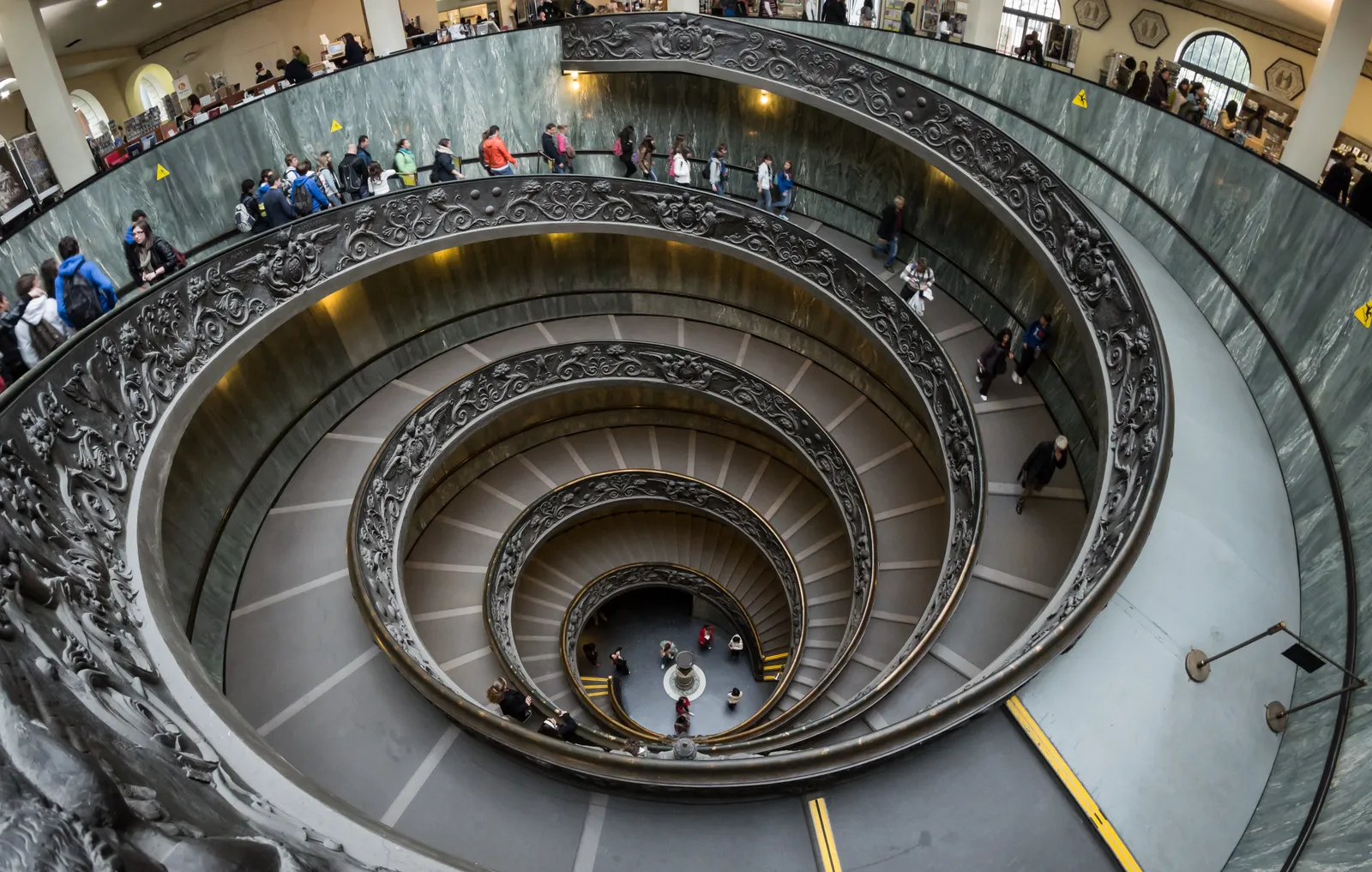 The double spiral staircase at the Vatican Museums seen from above, its bronze balustrade winding down through several turns to the floor far below, with visitors on the ramp
