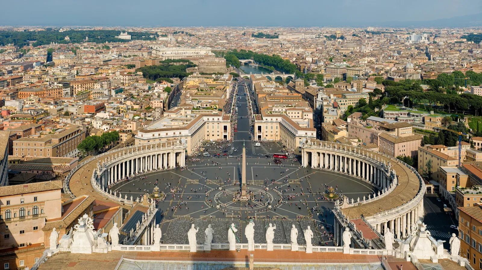 The view down from the top of St. Peter's dome over the whole elliptical square, both arms of the colonnade, the obelisk in the middle and the avenue running east toward the Tiber, with statues along the basilica roofline in the foreground