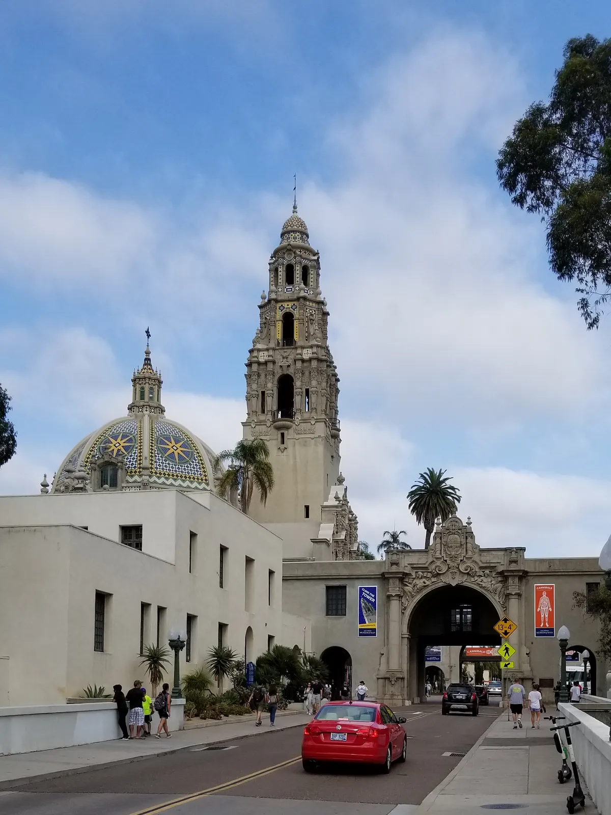 The California Tower and the blue-and-yellow tiled dome of the Museum of Us in Balboa Park, seen from the road running under the carved archway below them