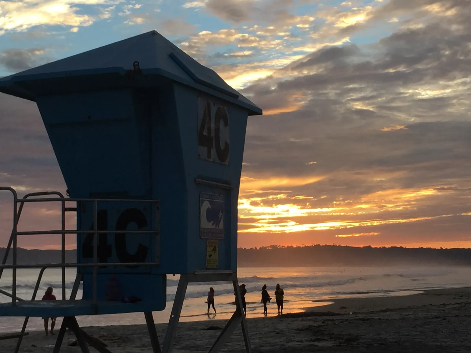 A blue lifeguard tower on Coronado Beach at sunset, with a handful of people walking at the waterline and cloud lit orange over the Pacific