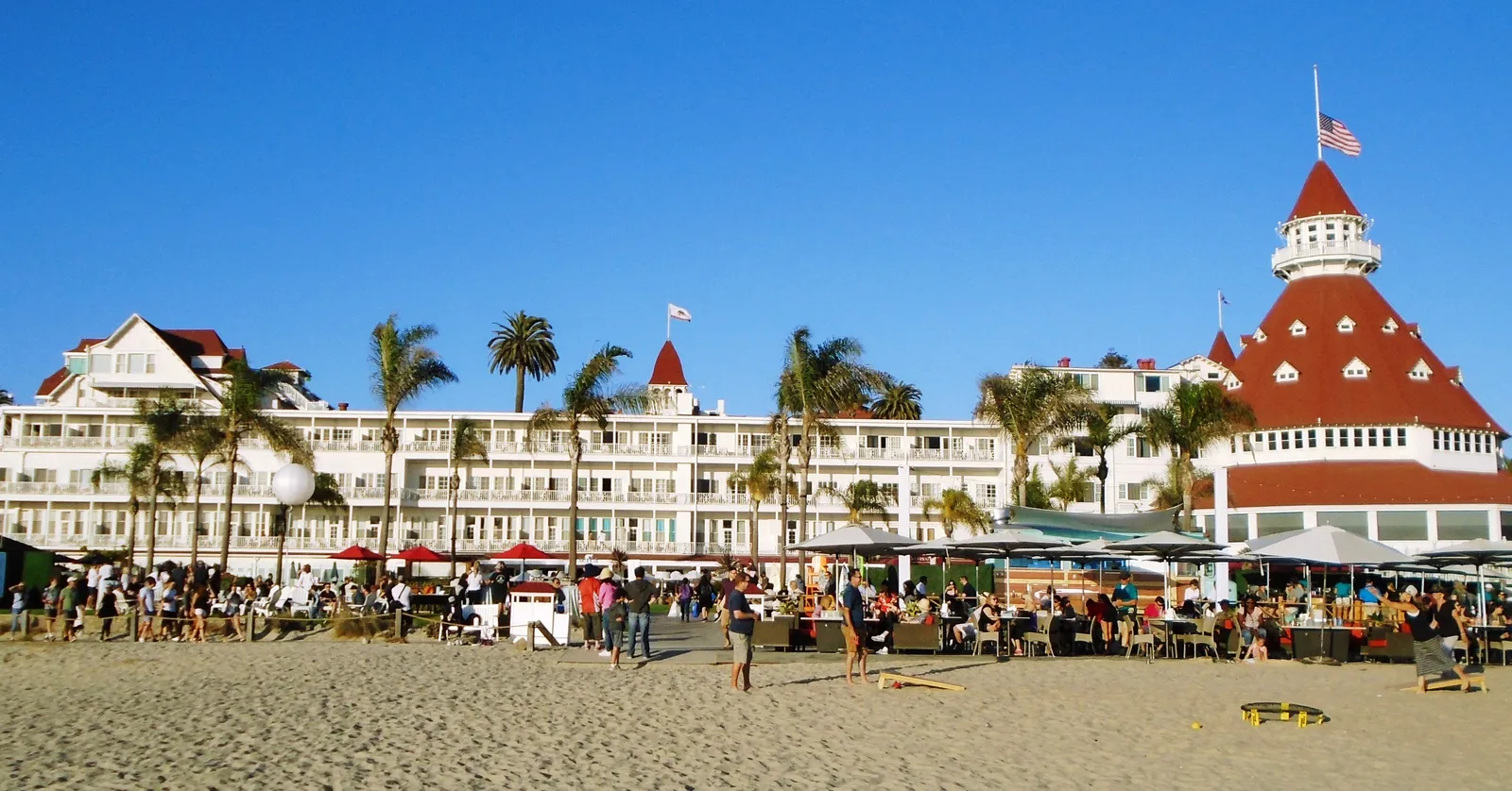 The white wooden Hotel del Coronado seen from the beach, with its red conical turret at one end, palms along the frontage and people on the sand below