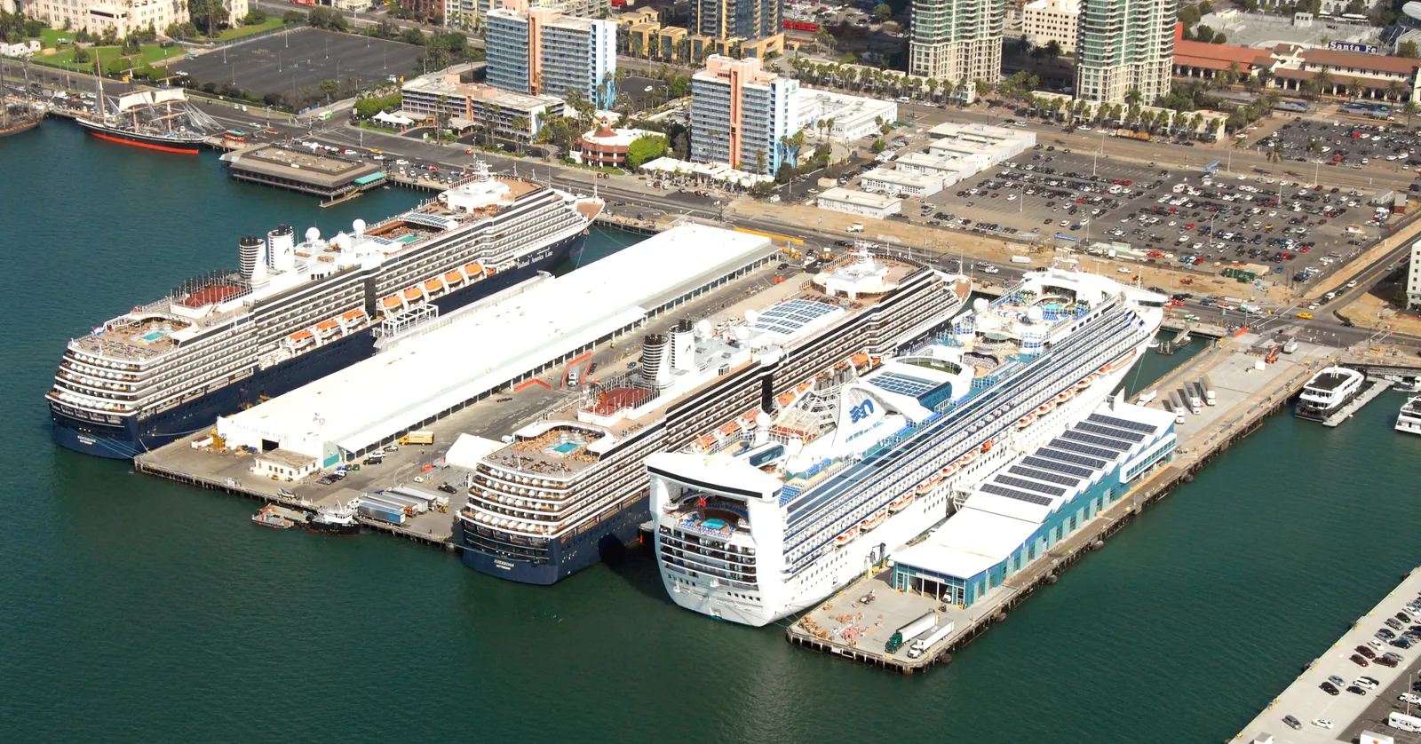 Three cruise ships tied up at the long white terminal building on B Street Pier in San Diego, seen from the air, with Harbor Drive and the downtown towers behind