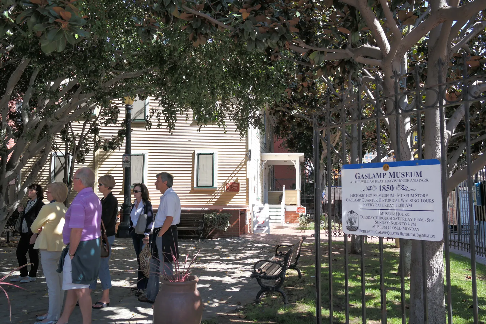 The pale wooden Davis-Horton House behind railings and shade trees in San Diego's Gaslamp Quarter, with a Gaslamp Museum sign at the gate and visitors reading it