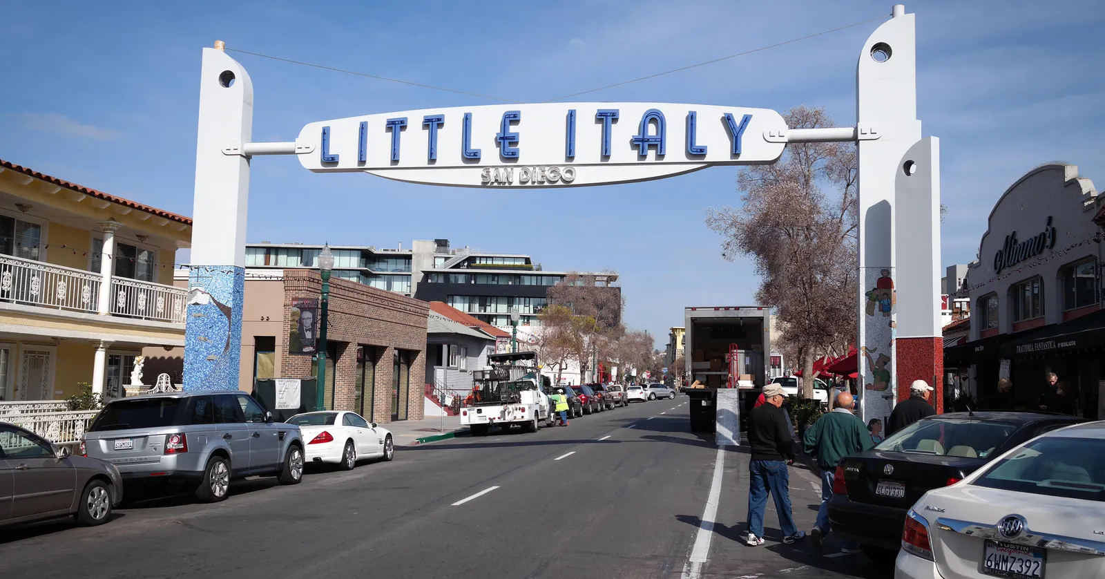 The white steel Little Italy sign arching over India Street in San Diego, with parked cars, low storefronts and palms along both pavements beneath it