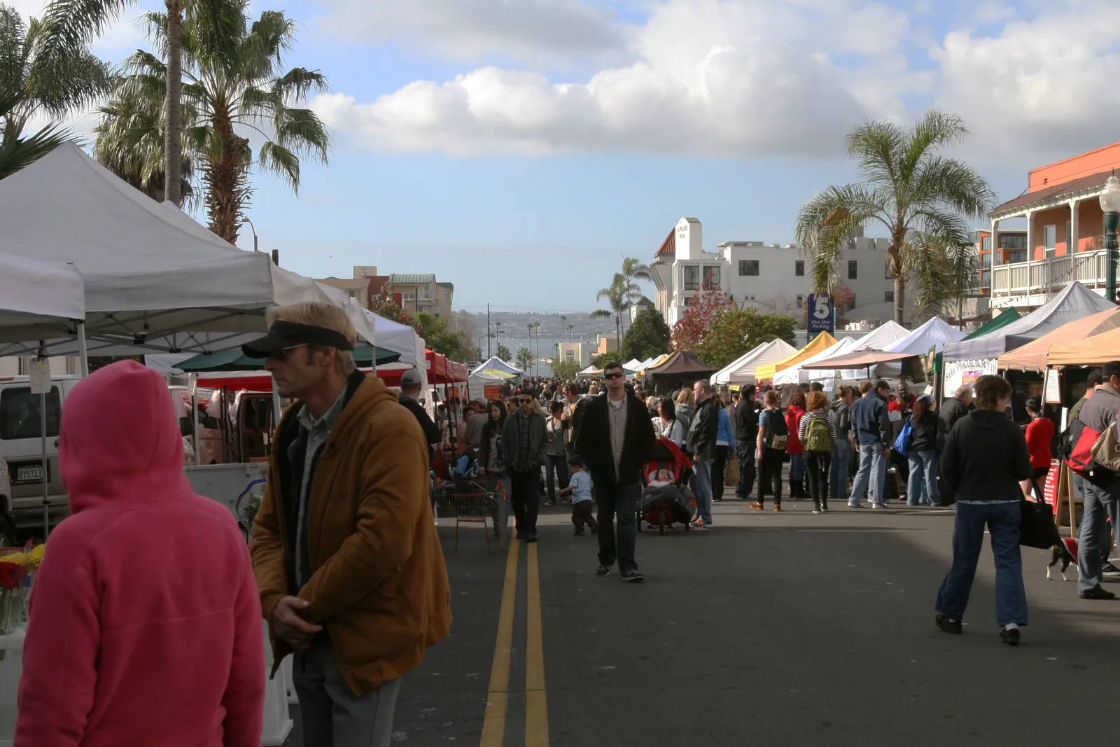 Crowds filling a closed street between rows of white market canopies at the Little Italy Mercato in San Diego, palms overhead and the harbor visible at the far end