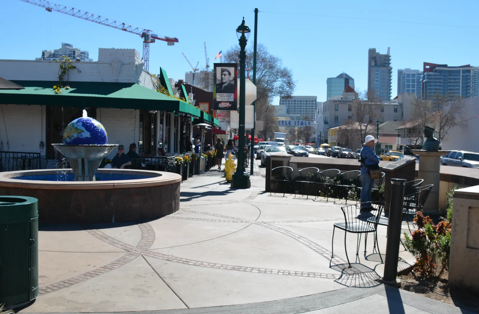 A round blue-tiled fountain on a paved piazza in San Diego's Little Italy, with café tables under awnings on one side and downtown towers in the distance