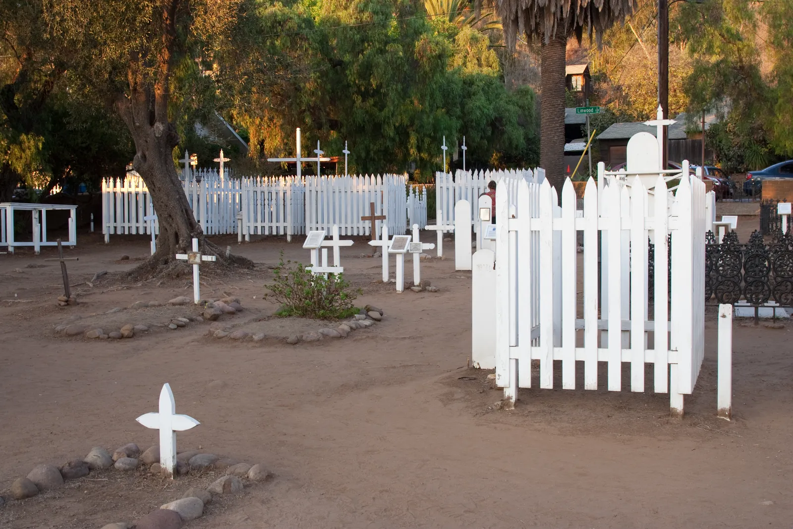 White wooden crosses and low picket fences on the bare earth of El Campo Santo cemetery in Old Town San Diego, under pepper trees at the end of the day