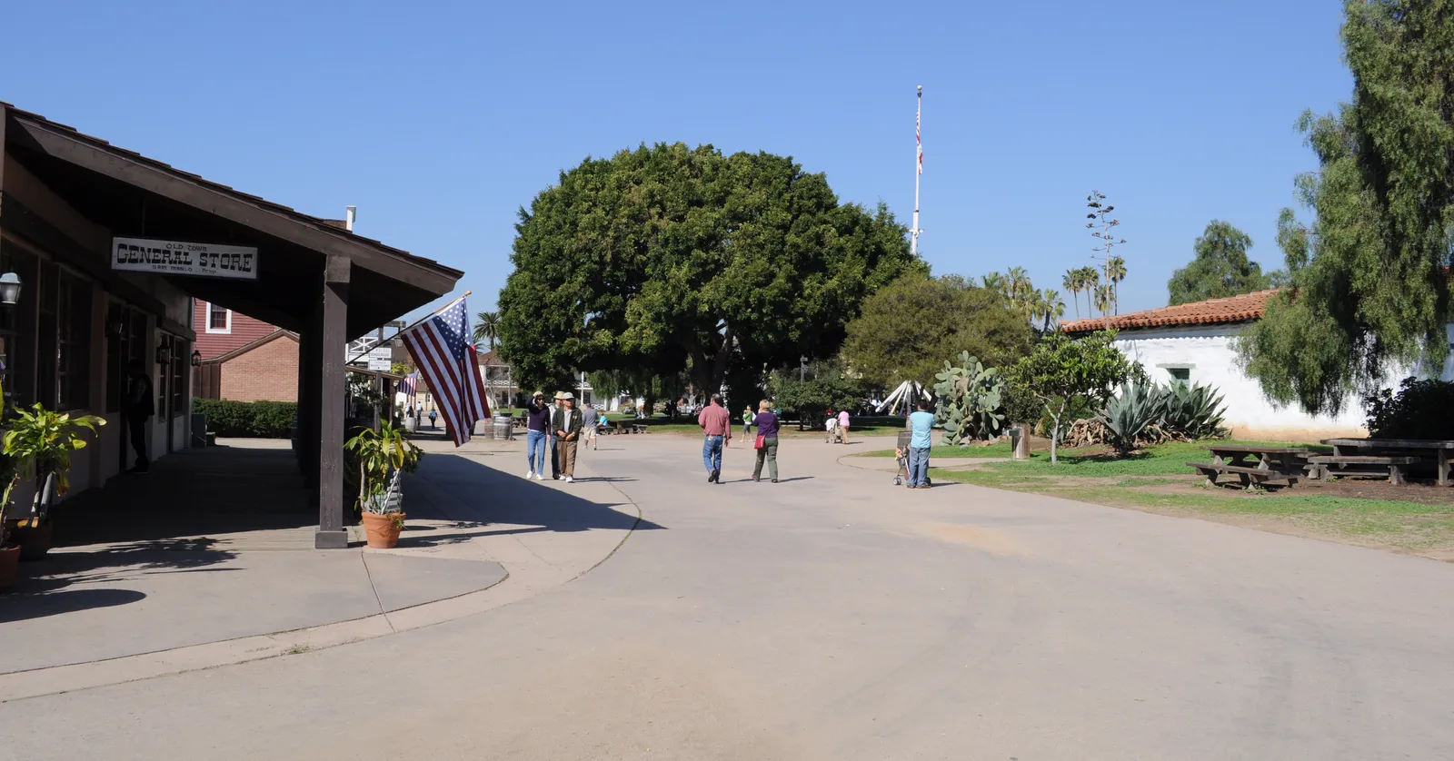 A packed-earth street through Old Town San Diego State Historic Park, with a wooden shop veranda on the left, a large shade tree in the middle and low white adobe buildings beyond