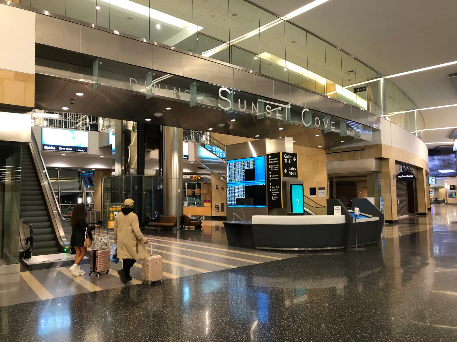 The lit Sunset Cove sign over the entrance to the food hall inside Terminal 2 at San Diego International Airport, with an escalator and travelers wheeling bags past