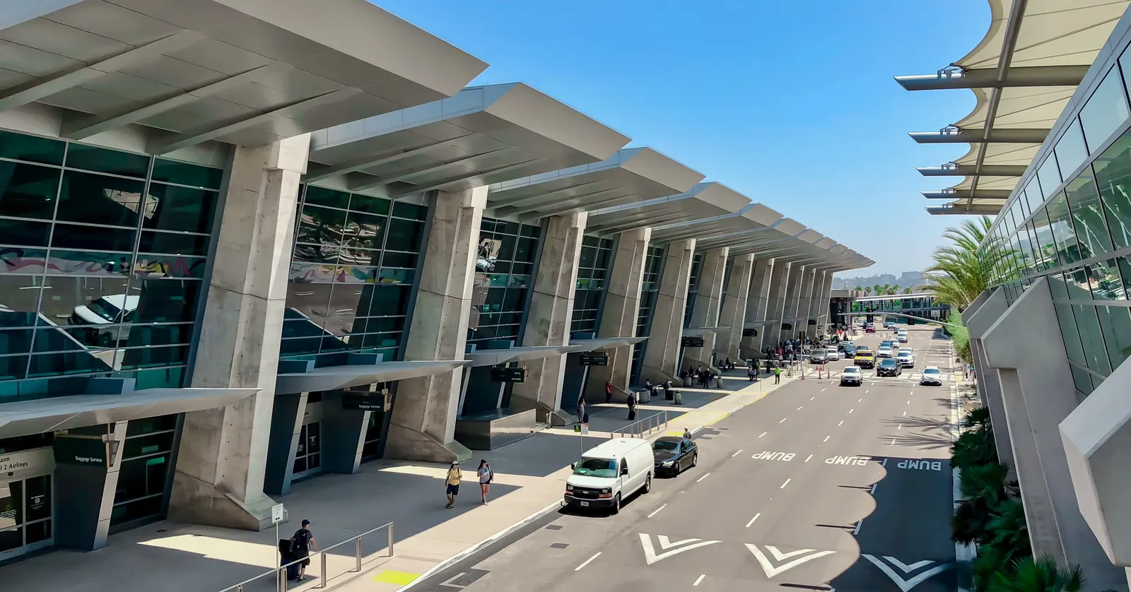 The curbside frontage of Terminal 2 at San Diego International Airport, its row of angled white canopies running along the departures road under a clear sky
