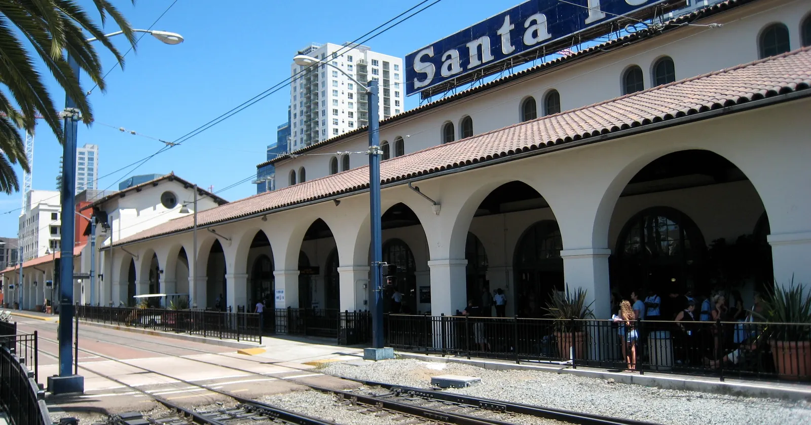 The arcaded front of Santa Fe Depot in San Diego under a blue sky, with the blue-and-white Santa Fe sign along the roofline and trolley tracks running past