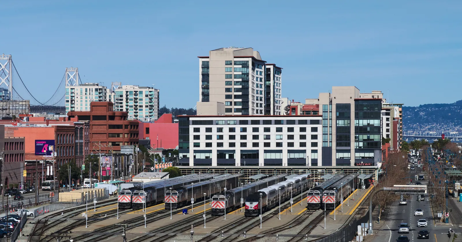 San Francisco's Caltrain terminus at 4th and King seen from above, red and white electric trains standing at the open platforms with SoMa apartment blocks and the Bay Bridge behind