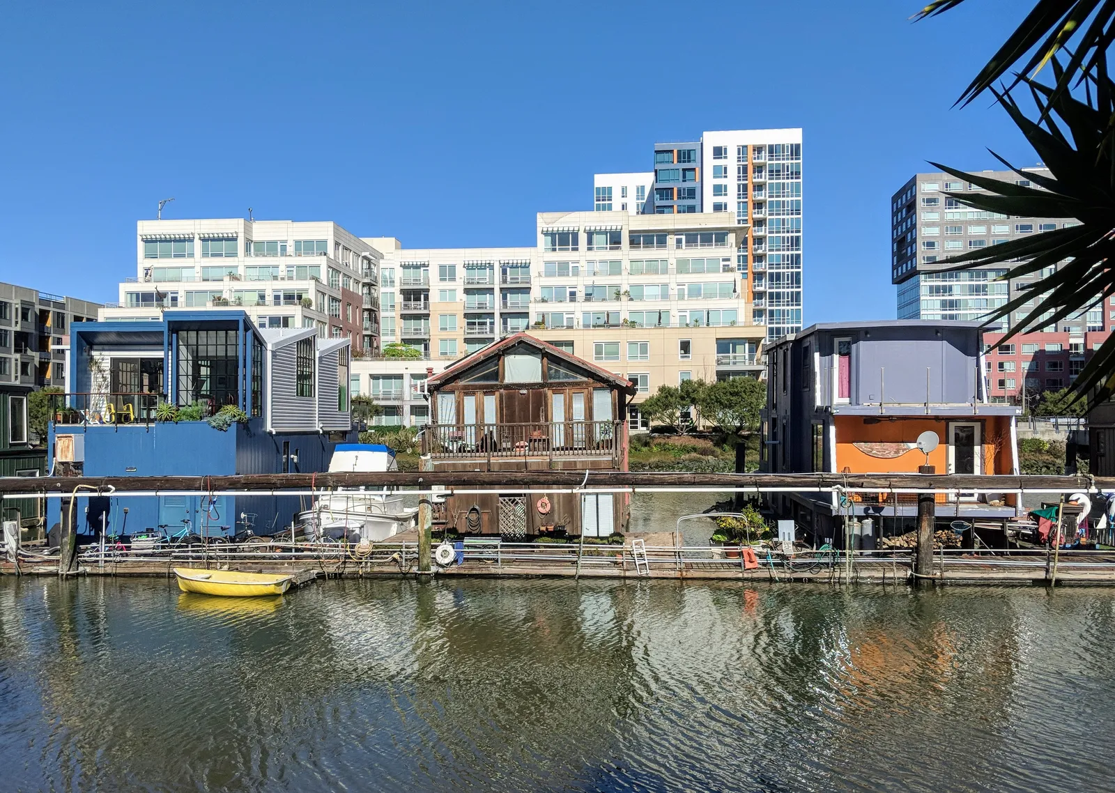 Houseboats moored along Mission Creek in San Francisco, a row of low timber and painted-metal homes on the water with glass apartment towers rising directly behind them