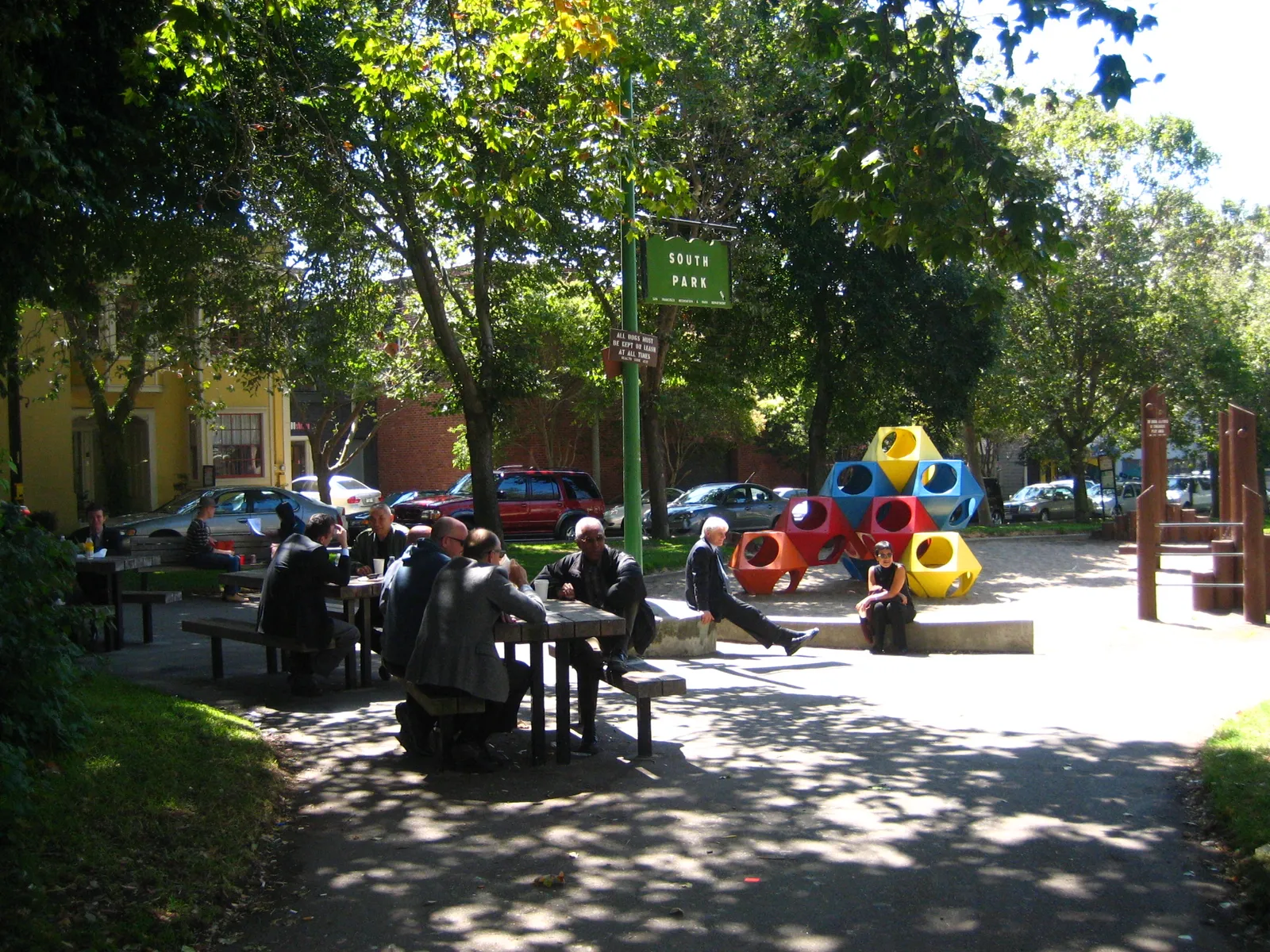 South Park in San Francisco, an oval of grass and mature trees with people sitting at picnic tables, a play structure to one side and low brick buildings around the edge