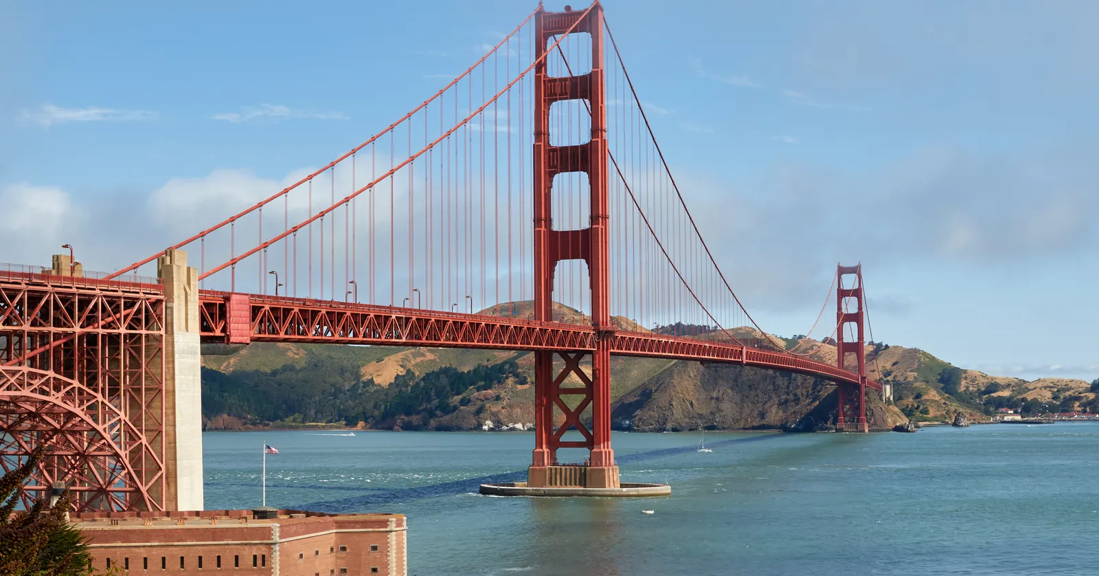 The Golden Gate Bridge in bright sun from the San Francisco side, both orange towers and the full suspended span crossing the strait with the green Marin headlands rising behind