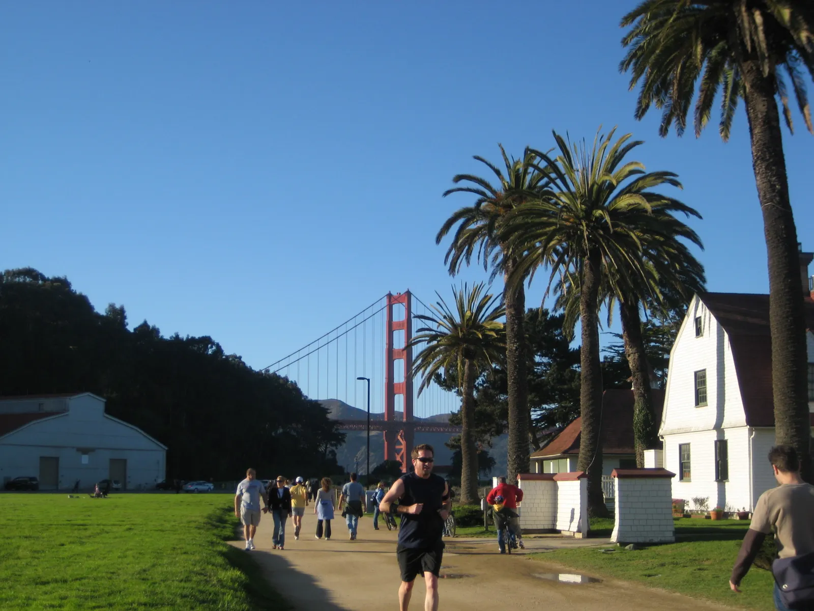 The gravel promenade at Crissy Field in San Francisco, walkers and a runner passing under tall palms beside a white clapboard building with the Golden Gate Bridge tower in the distance