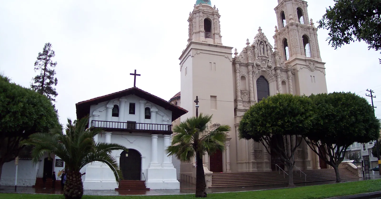 The small whitewashed adobe chapel of Mission Dolores in San Francisco standing beside the much larger ornate stone basilica of 1918, with palms and street trees in front of both