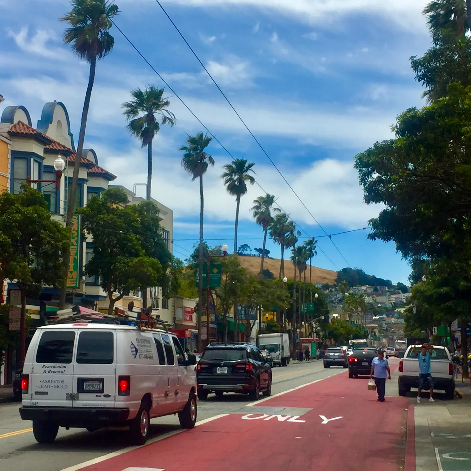 Mission Street at 24th Street in San Francisco, palm trees and low shopfronts along a wide sunny street with a red transit lane painted down the middle and hills in the distance