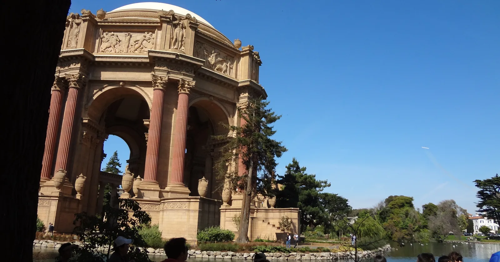 The rotunda of the Palace of Fine Arts in San Francisco rising in ochre stone-coloured concrete above its lagoon, with the curved colonnade and cypress trees running away to the right