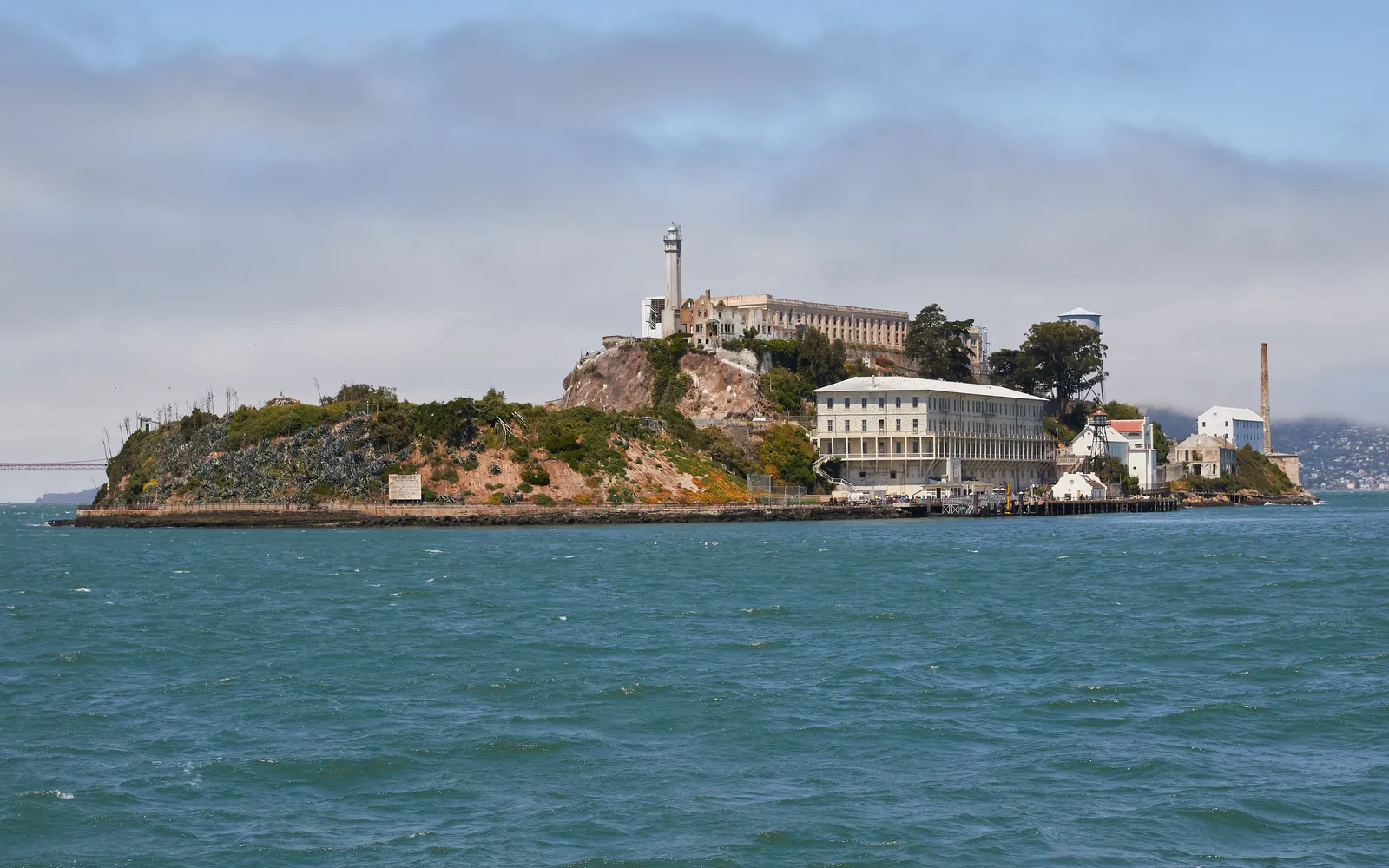 Alcatraz Island in San Francisco Bay seen from the water, with the long cellhouse and lighthouse on top of the rock and the old barracks building at the waterline