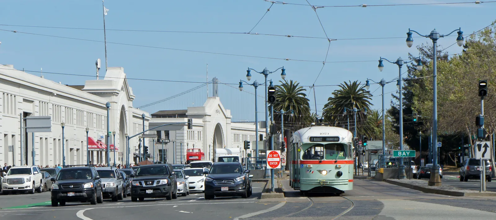 A green and cream vintage streetcar on the F line running along the Embarcadero in San Francisco, past the arched fronts of the old pier sheds