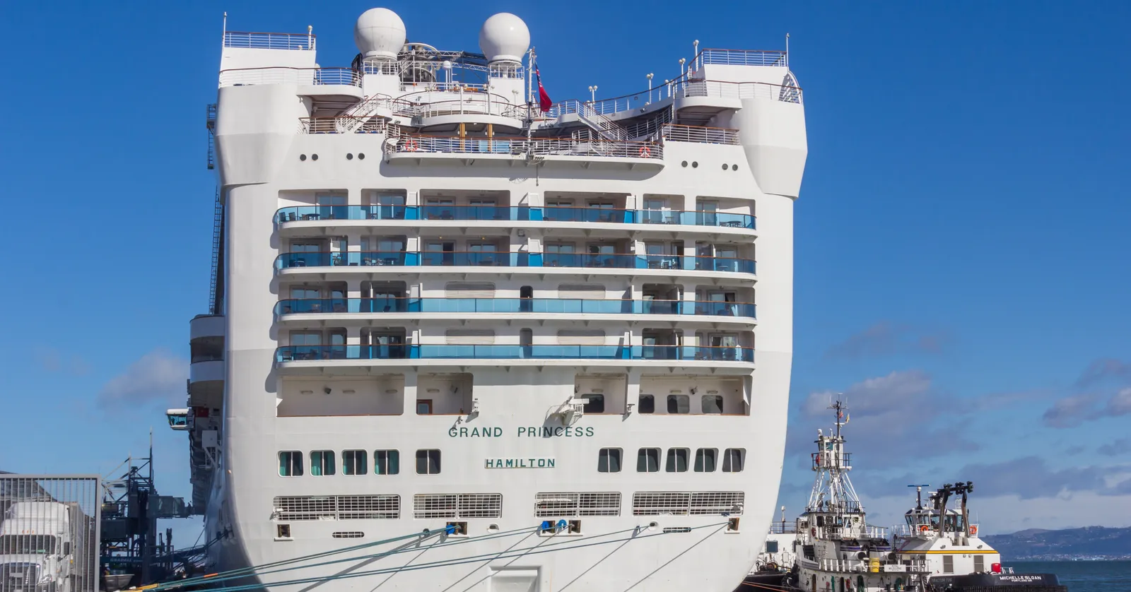 The white stern of a cruise ship tied up at Pier 27 in San Francisco, with balconies stacked across it and two tugboats waiting alongside under a blue sky