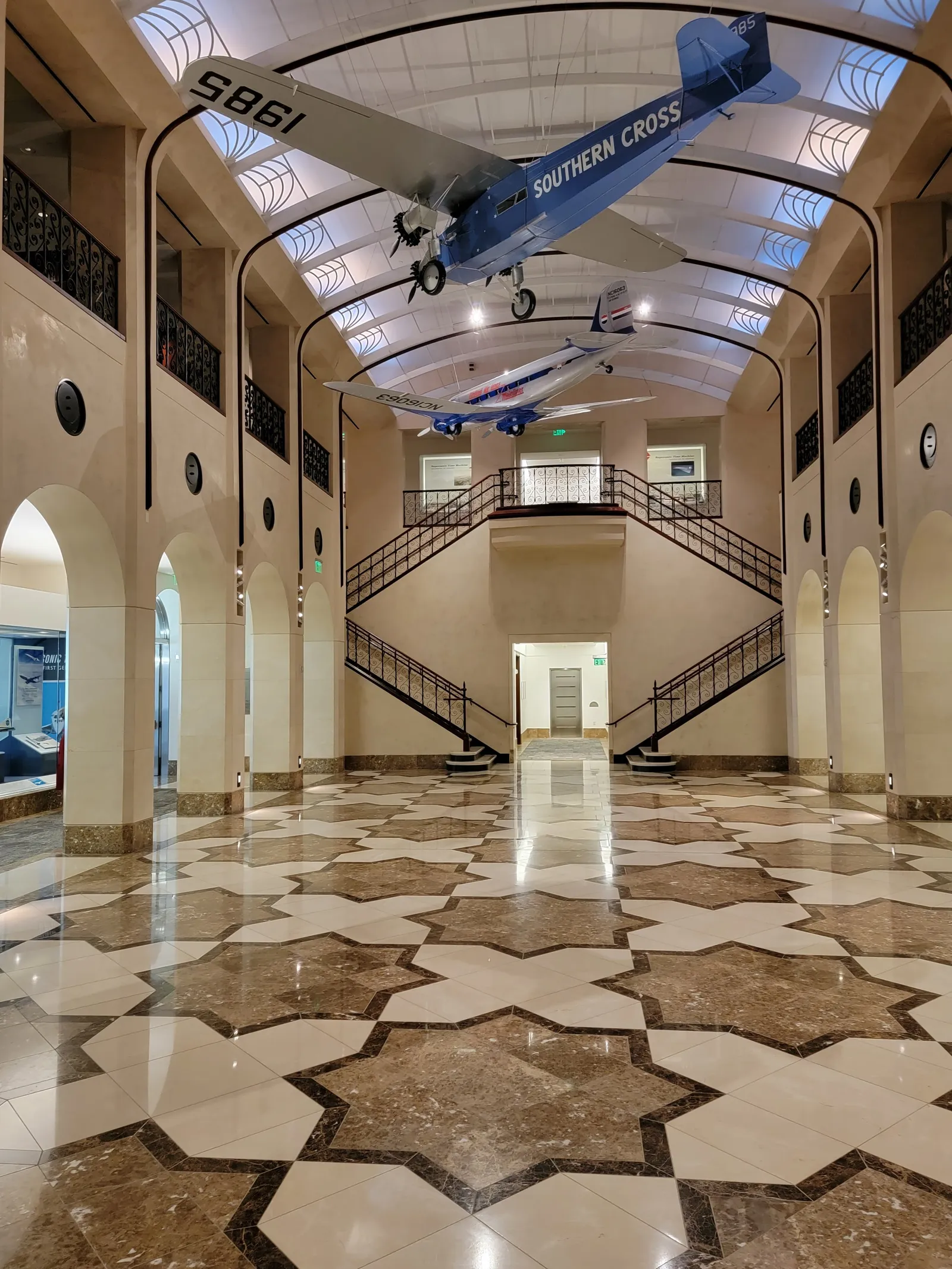 The main room of the aviation museum at San Francisco International Airport, an arcaded hall with a starred marble floor, twin staircases at the far end and a model of the Southern Cross aircraft hanging from the barrel-vaulted ceiling