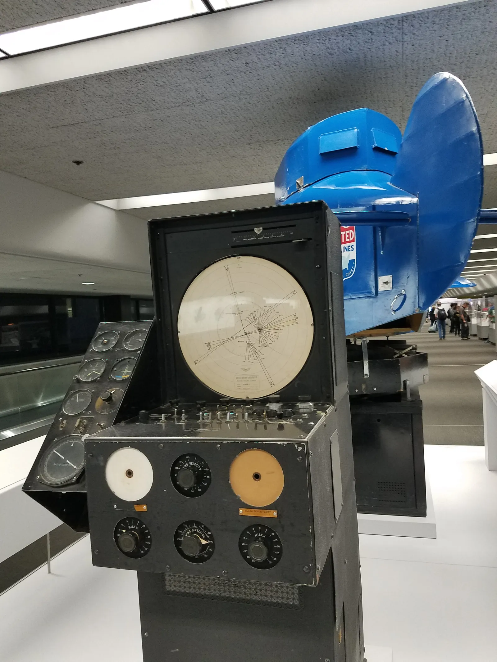 A blue Link flight trainer from the 1940s and its instructor console on display in a glass-walled airport concourse at San Francisco International Airport, with an escalator and passengers behind it