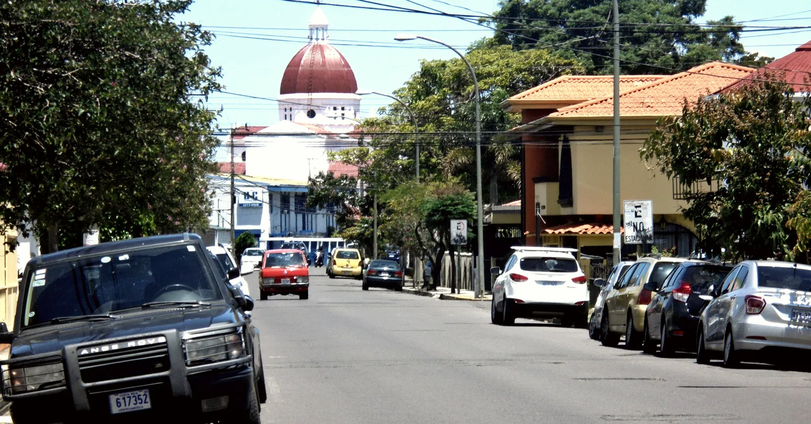 A residential street in Barrio Escalante, San Jose, cars parked along both kerbs under tiled roofs and street trees, with the red dome of a white church closing the view at the far end