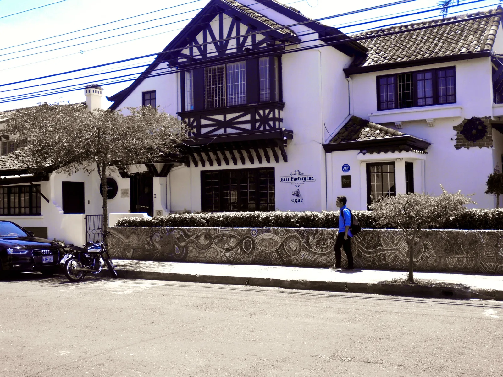 A white house in Barrio Escalante with dark mock-Tudor timbering in its gable and a low garden wall covered in coloured mosaic, a motorbike parked in front and a man walking past