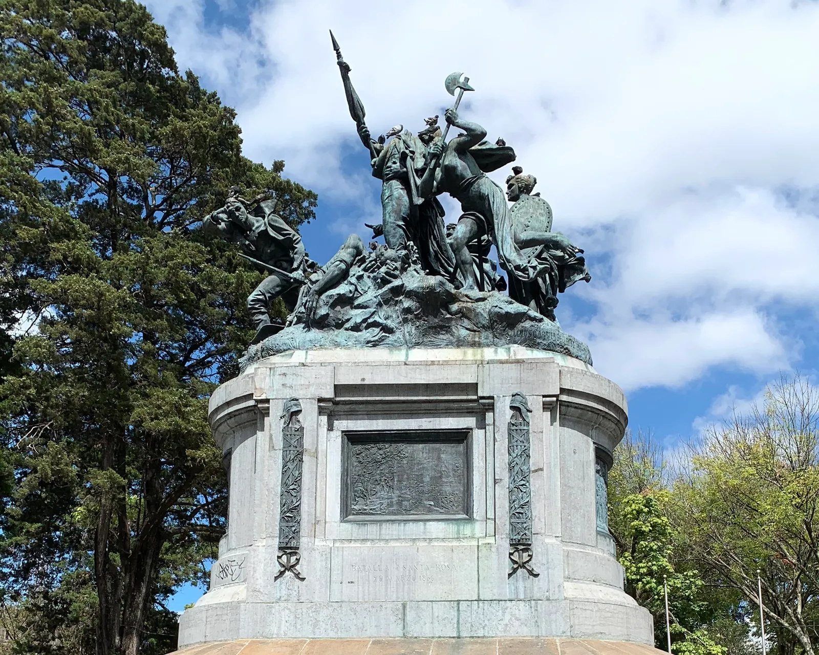 The bronze group of the National Monument in San Jose on its tall stone drum, several figures pressing forward around a standard, with dark cypress trees behind