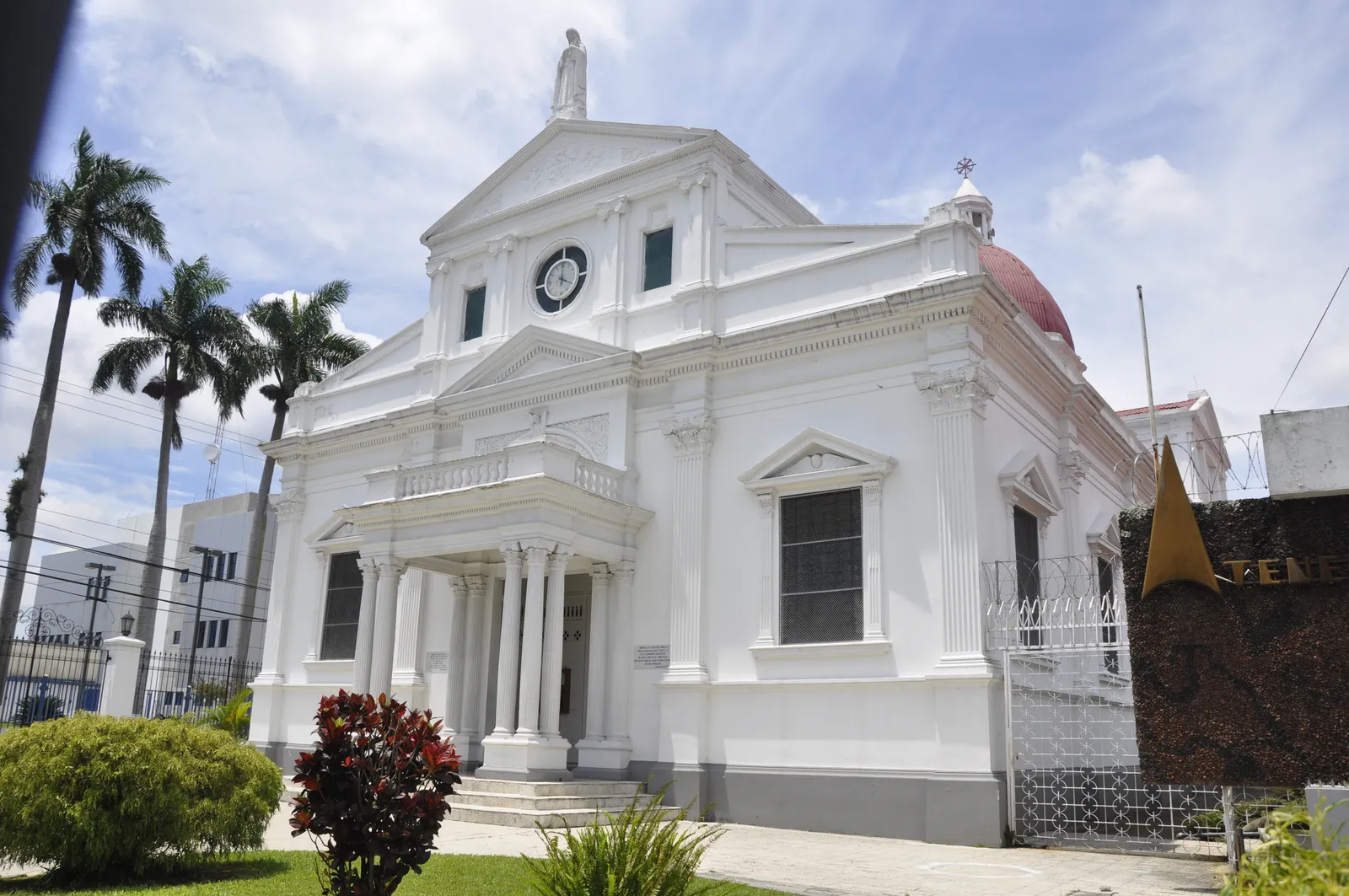 The white front of the Santa Teresita church in San Jose, a columned porch under a triangular pediment with a clock above it, a statue on the roofline and the red dome behind