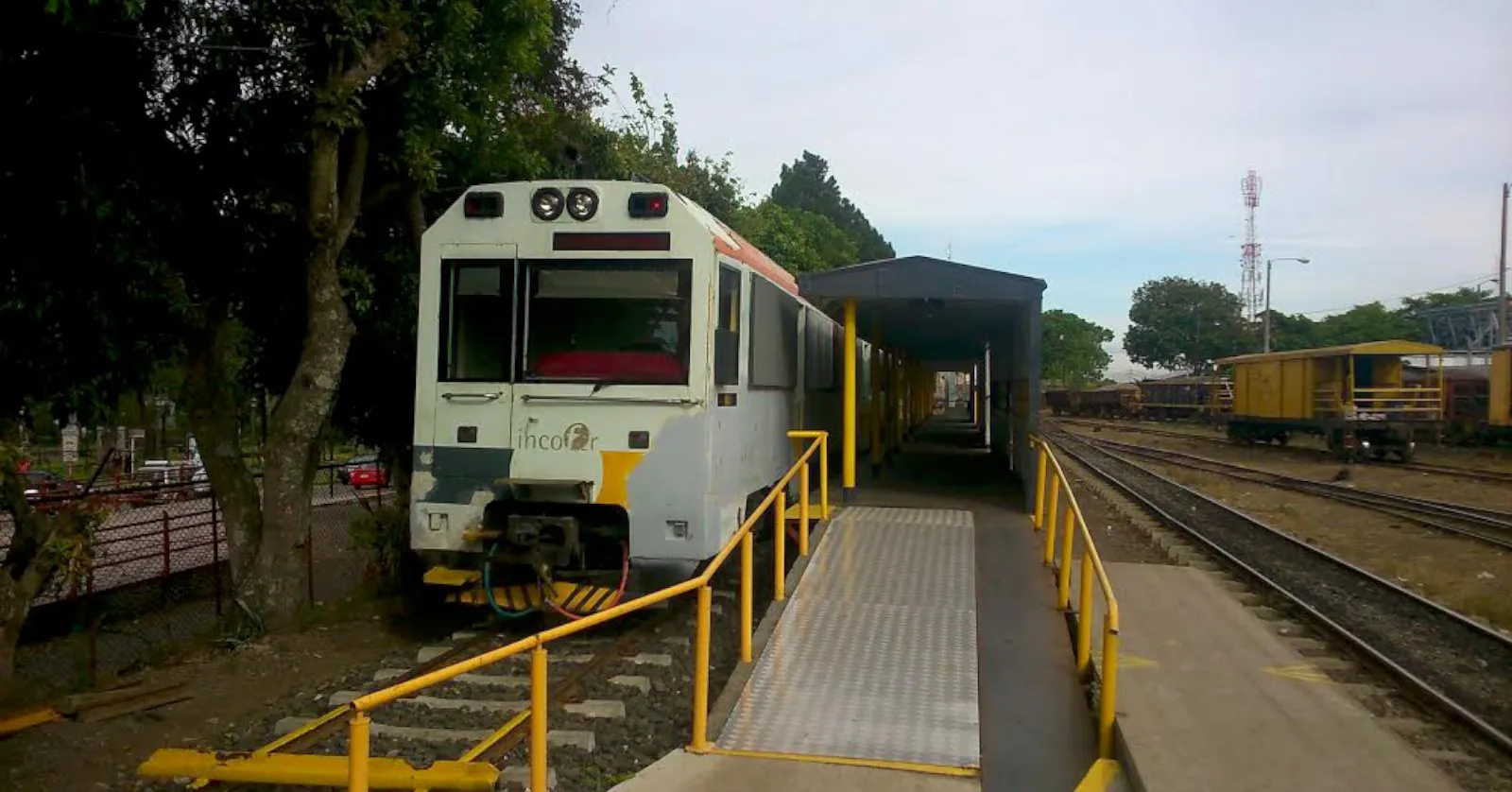 A white and yellow diesel railcar standing at the bay platform of Estación al Atlántico in San Jose, yellow handrails along the ramp beside it and freight wagons on the tracks to the right