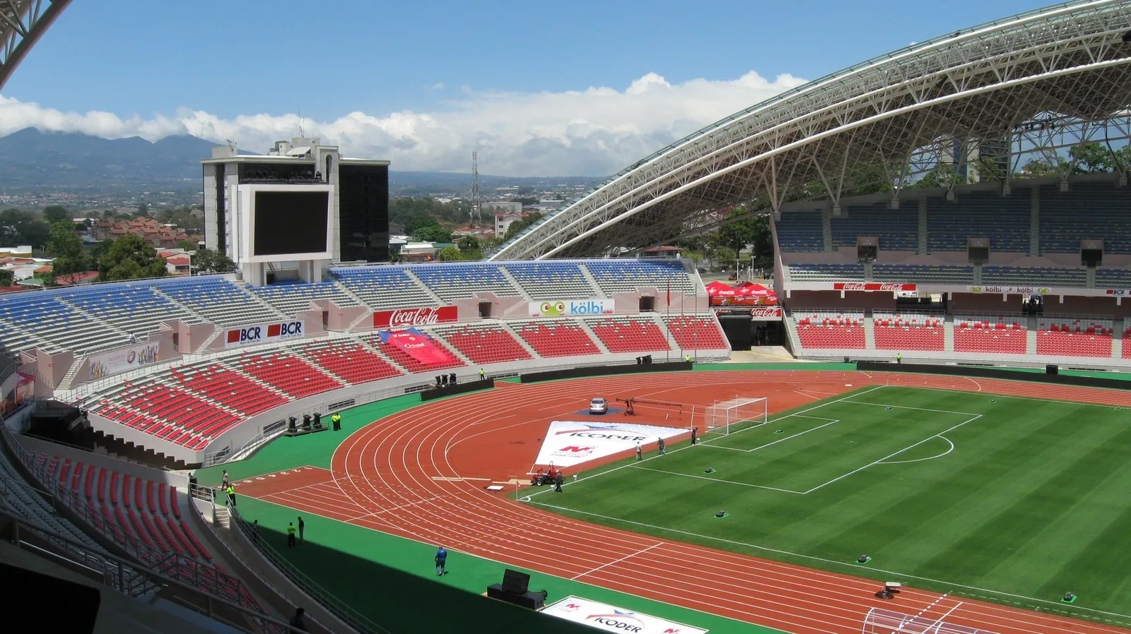 Inside Costa Rica's National Stadium, red and blue seats under a curved lattice roof, an orange running track around the pitch and green mountains beyond the far stand