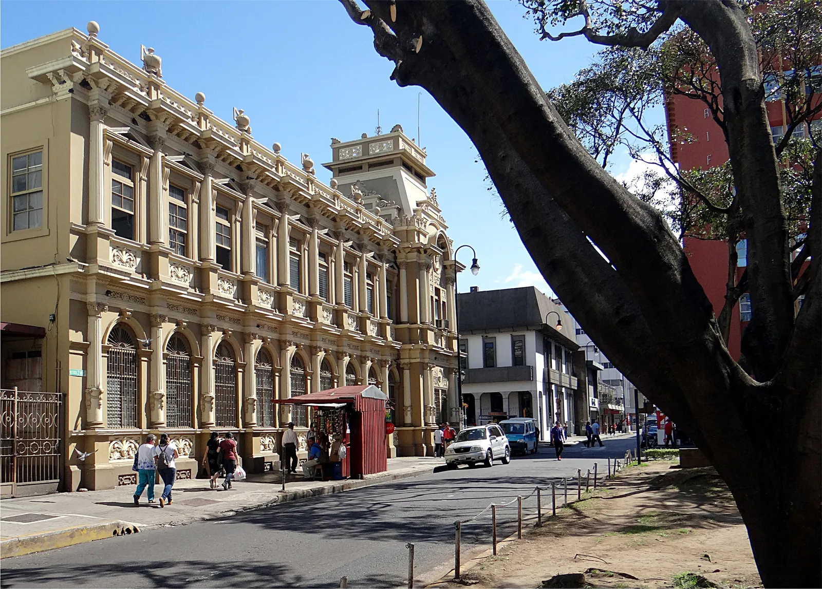 The pale stone front of San Jose's central post office, a long arcaded facade with balustrades and a tower at the far corner, seen along the street from under a tree