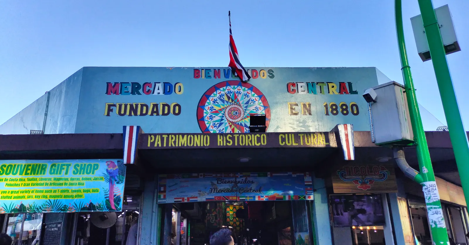 A painted teal entrance front to San Jose's central market reading Bienvenidos, Mercado Central, fundado en 1880, patrimonio historico cultural, with a Costa Rican flag above it and shop signs underneath