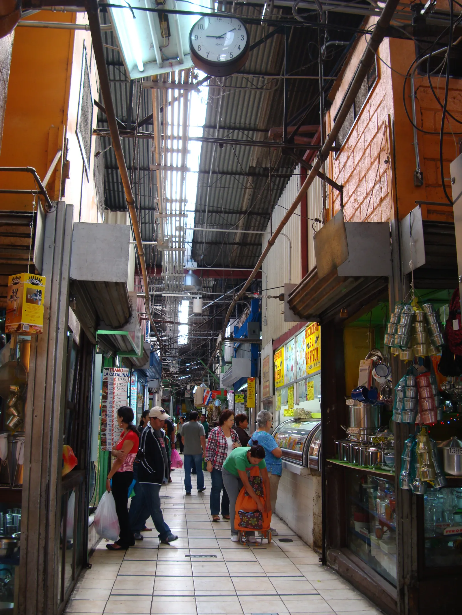 A narrow tiled aisle inside San Jose's central market running away under a corrugated roof hung with cables and a clock, stalls trading on both sides and shoppers passing between them