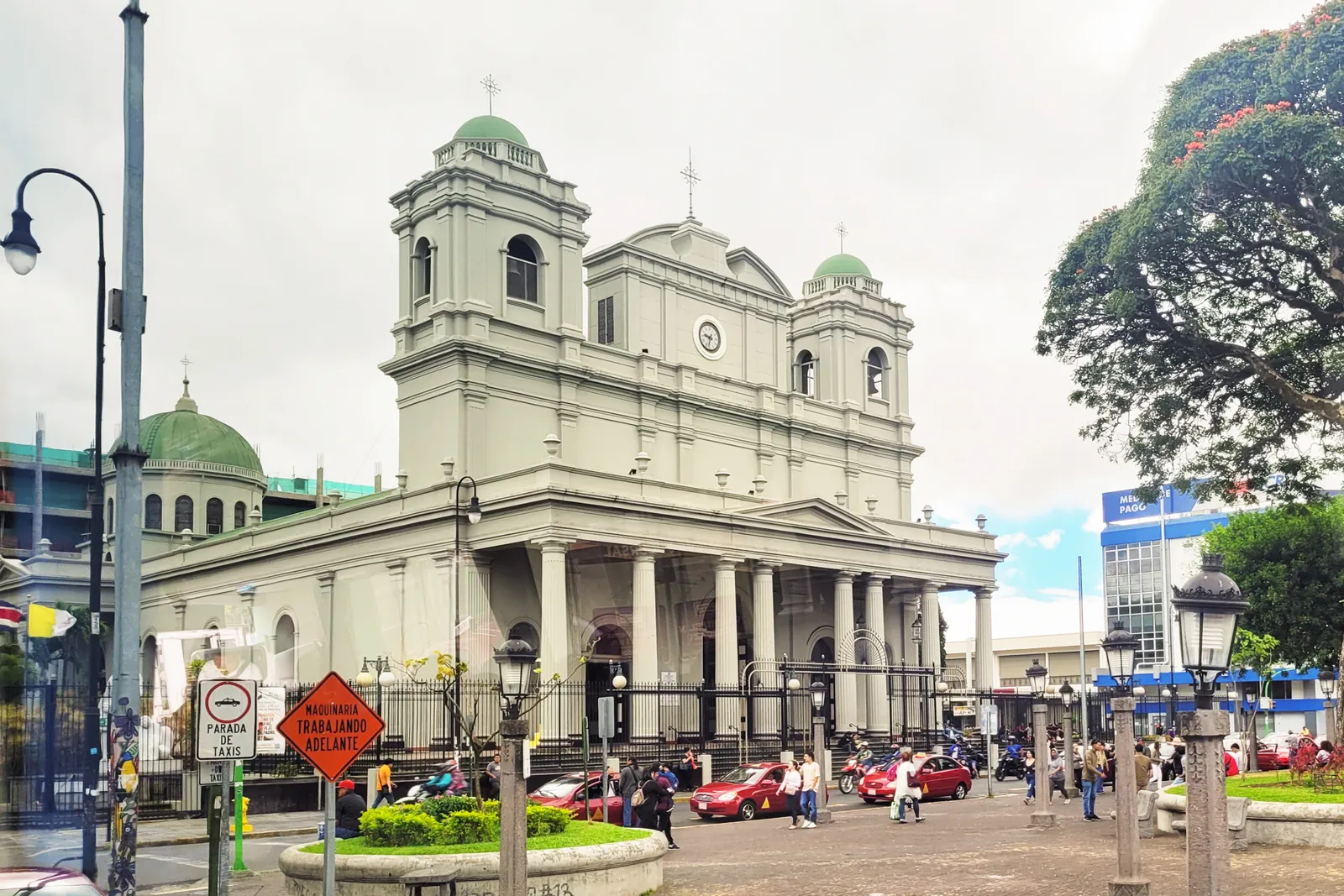 The Metropolitan Cathedral of San Jose seen across Parque Central, a grey-white front with a columned portico, a clock in the gable and green copper domes behind