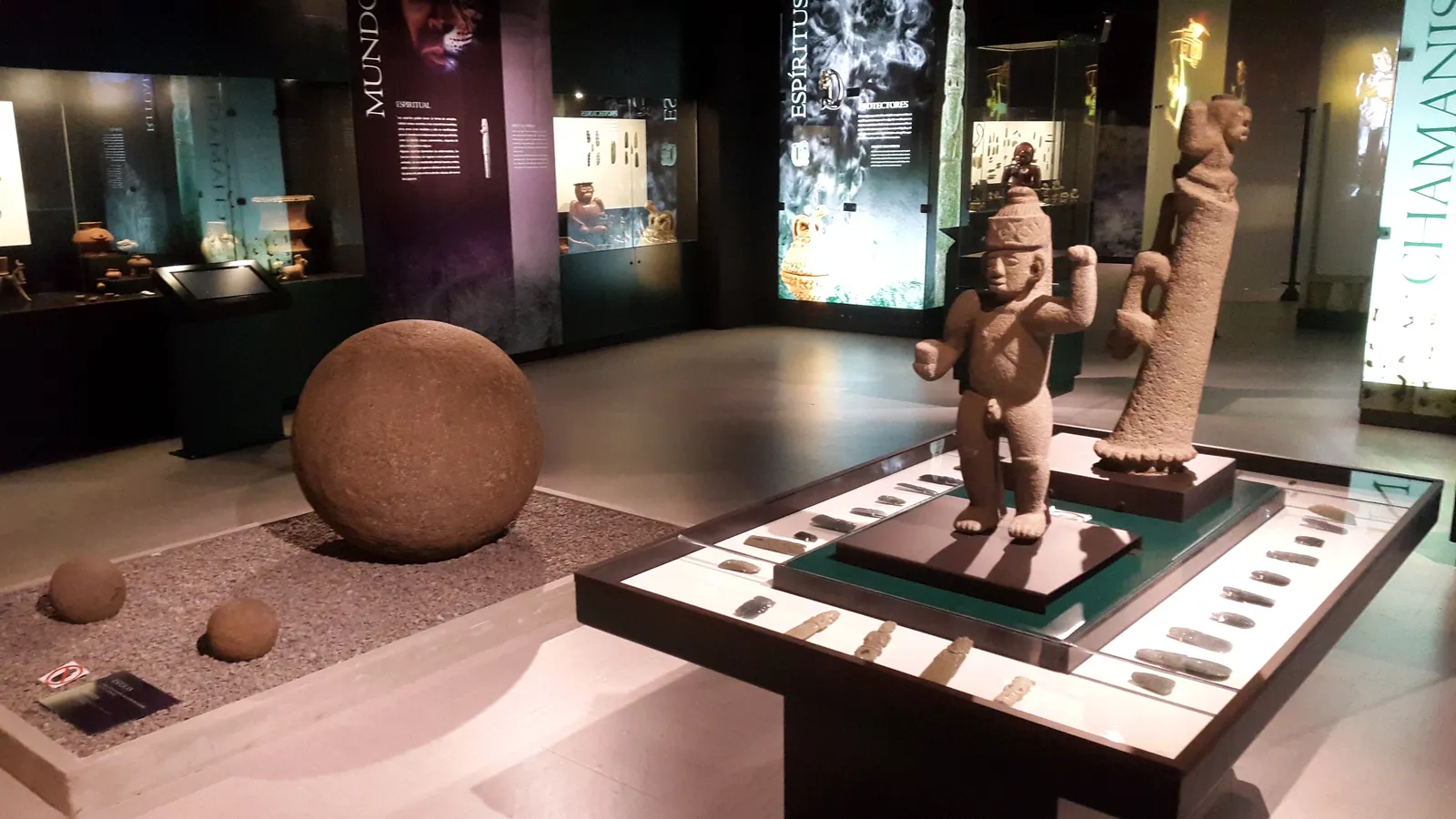 A spotlit gallery in San Jose's jade museum with a carved stone sphere on gravel, a standing stone figure on a plinth and lit wall panels behind