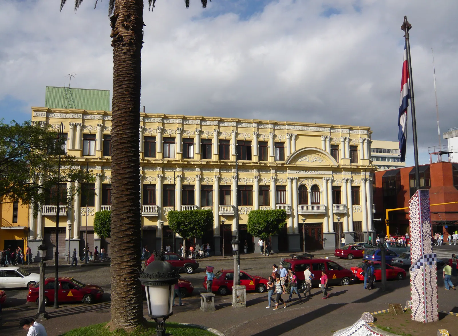 The pale yellow two-storey front of the Melico Salazar theatre in San Jose behind a palm tree, with red taxis parked along the street and a Costa Rican flag flying at the corner