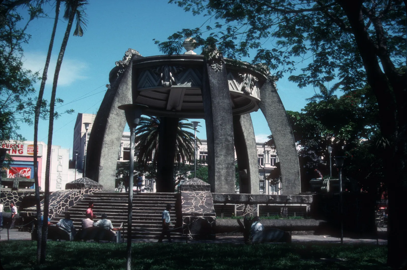 The concrete bandstand in San Jose's Parque Central, six curved pillars leaning in to carry a round upper platform, with steps up to it and palms on either side