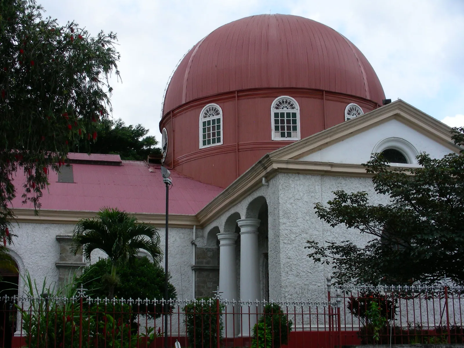 The deep red dome of Alajuela's cathedral rising behind a white portico of columns, with palms and a railing in front