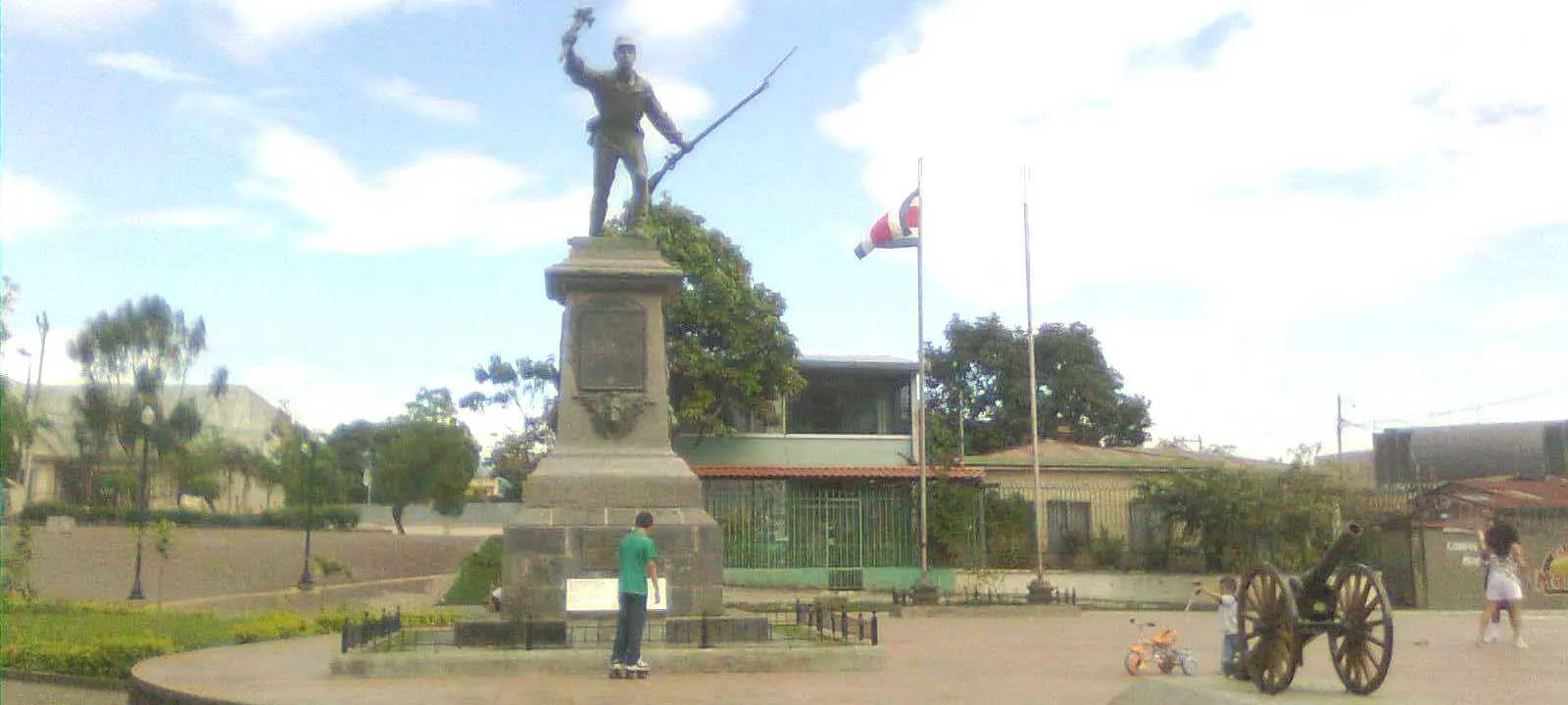 The bronze figure of Juan Santamaría on its tall stone pedestal in the middle of a paved Alajuela square, with a cannon on the paving to the right and a Costa Rican flag behind