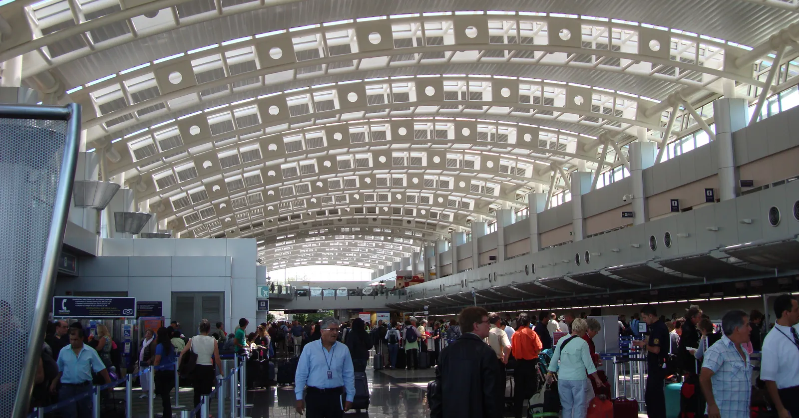 Passengers with rolling suitcases queueing under the curved white lattice roof of the check-in hall at Juan Santamaría International Airport