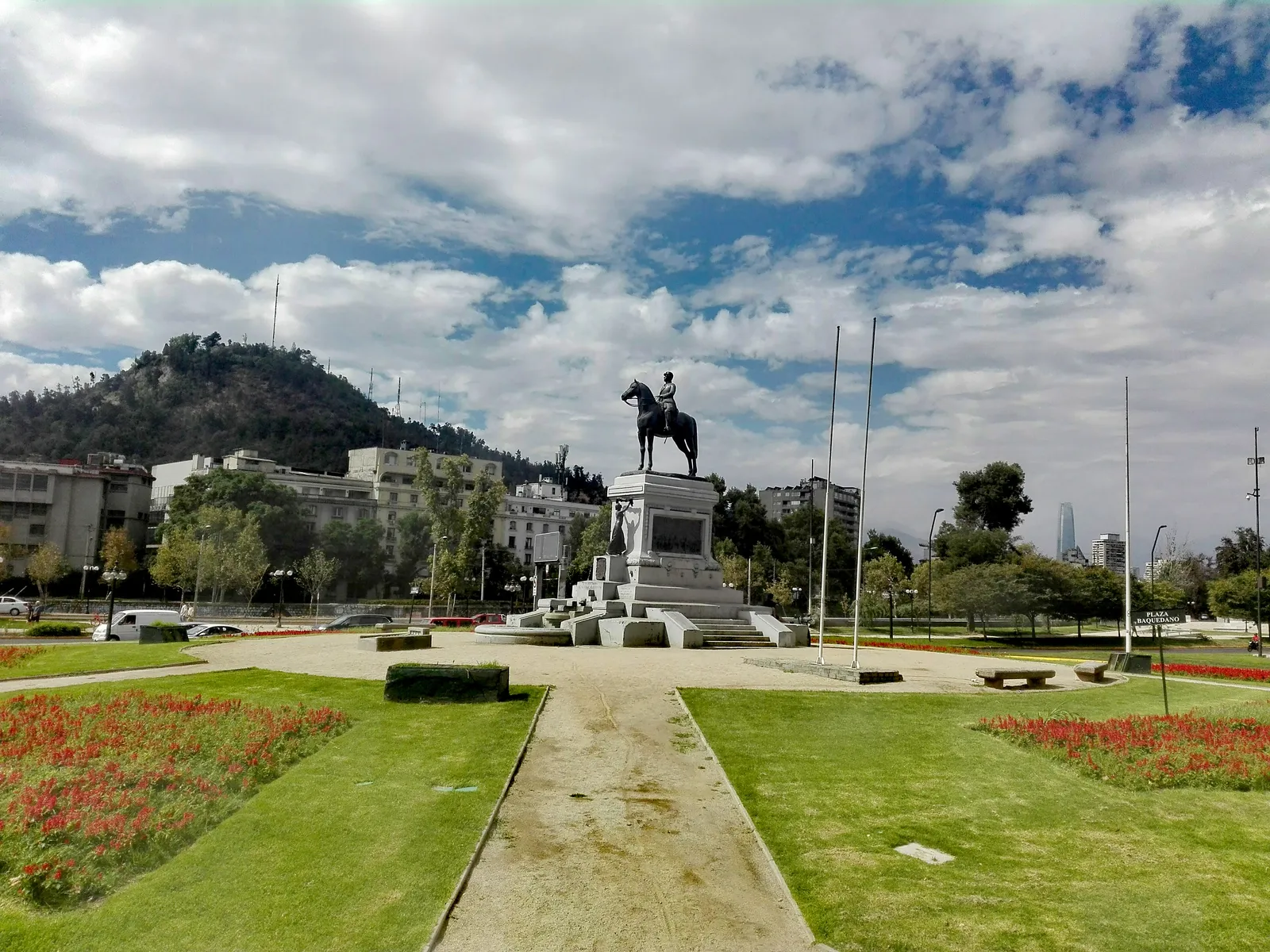 The bronze equestrian statue of General Baquedano on its stone pedestal in the middle of Plaza Baquedano, Santiago, with lawns, flowerbeds and a wooded hill behind