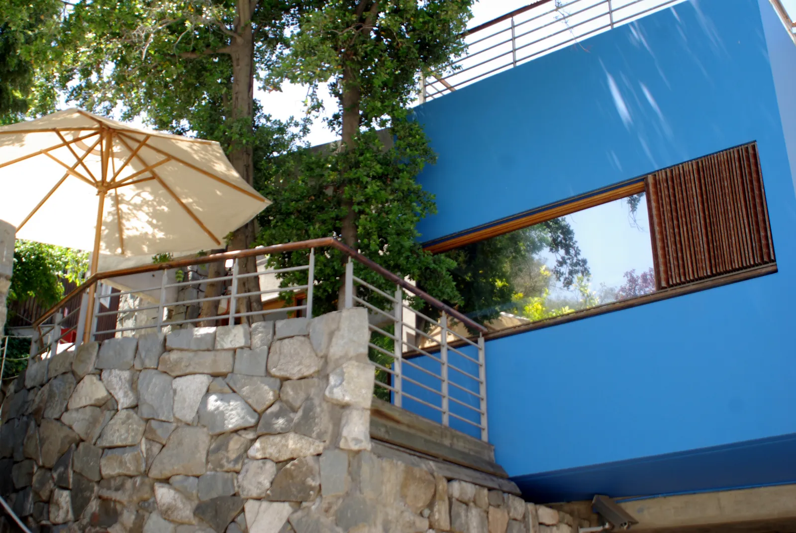 The bright blue upper storey of Pablo Neruda's house La Chascona in Santiago, above a rough stone retaining wall and a shaded terrace with a parasol