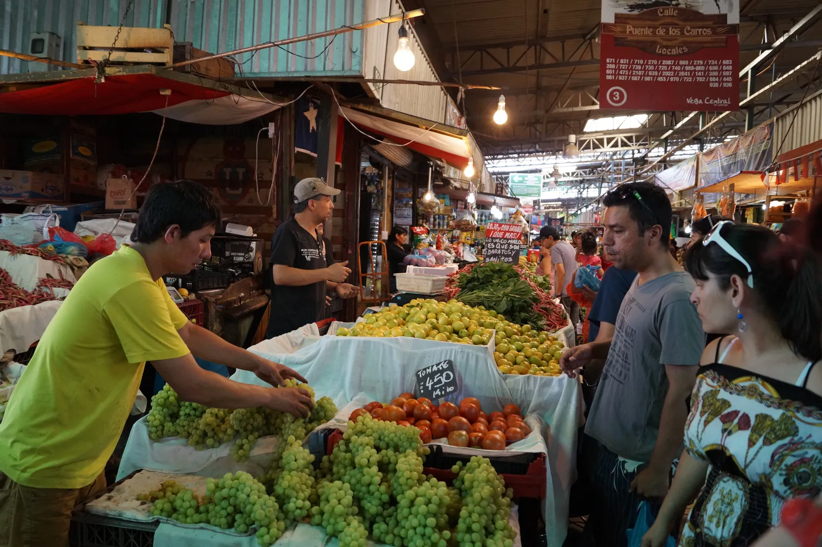 Stalls piled with grapes, tomatoes and other produce under a corrugated roof at La Vega Central market in Santiago, with traders and shoppers along the aisle