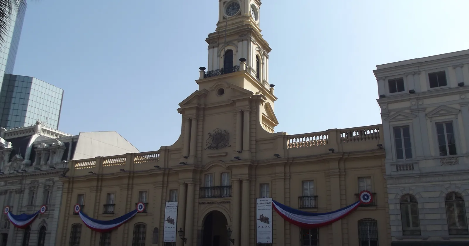 The pale yellow colonial facade and clock tower of the Palacio de la Real Audiencia on Santiago's Plaza de Armas, with Chilean flag bunting strung along its front