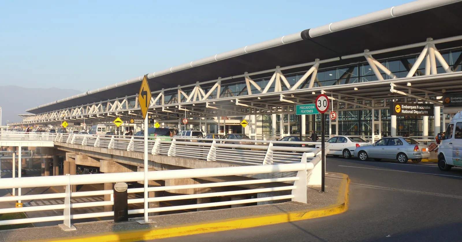 The kerbside frontage of Santiago's airport terminal, a long low canopy on angled steel supports with cars pulling in beneath it and mountains in the distance